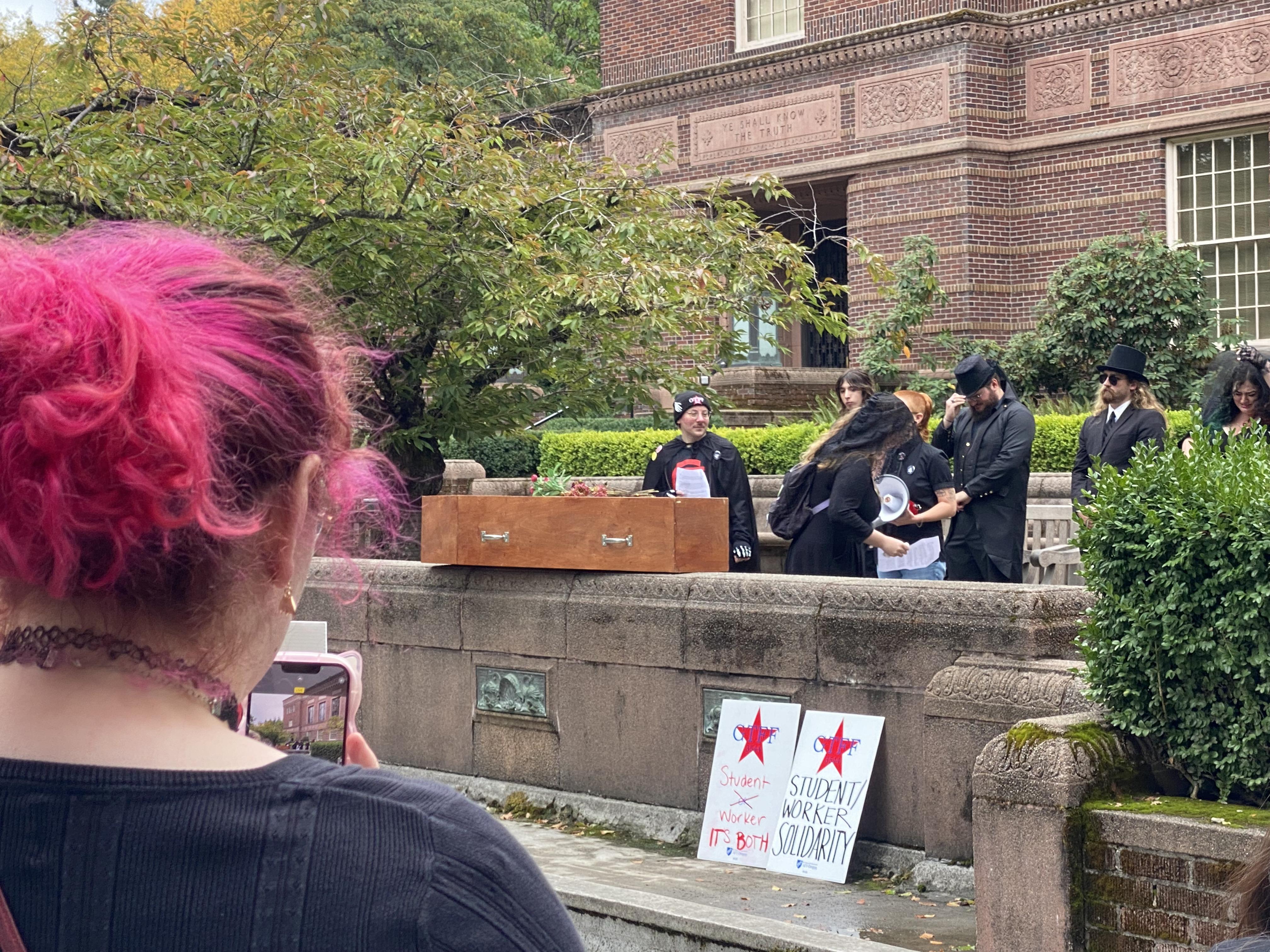 Members of the GTFF and UO Student Workers stand behind a wooden casket, dressed for a typical funeral wake. Multiple speeches were given, predominantly featuring testimony from graduate student workers about how their wages are incapable of covering the cost of living in Eugene. Two signs at the front read “Student or Worker[sic] It’s Both!” and “Student/Worker Solidarity”