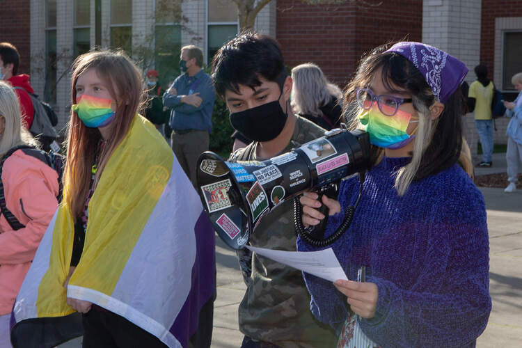 Photo of students protesting, with one of them using a nonbinary flag as a cape.
