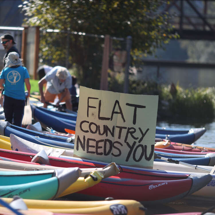 a sign reading &ldquo;Flat Country Needs You&rdquo; is surrounded by kayaks along the river shore
