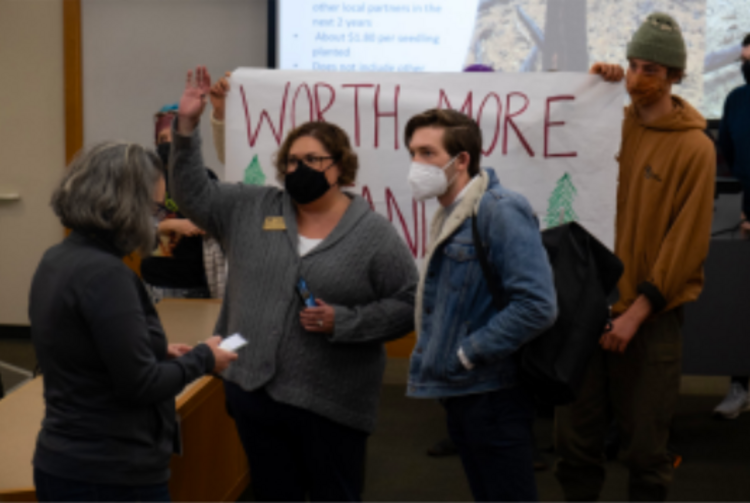 Protestors interrupt the presentation on post-fire logging, in front of a banner reading &ldquo;Worth More Standing&rdquo;