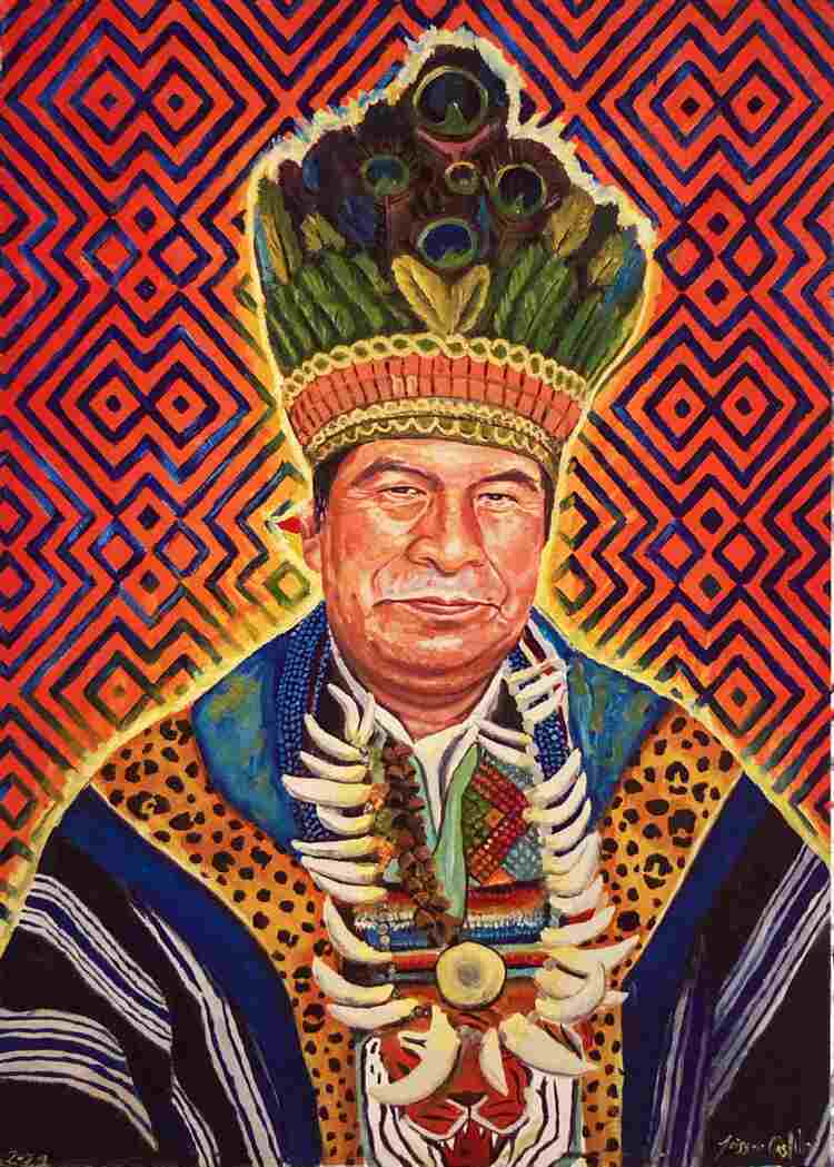 A Kamëntsá shaman in ceremonial regalia sits in front of a colorful background of geometric desings. He wears a woven tunic, necklaces of beads and jaguar teet, and a feather headdress.